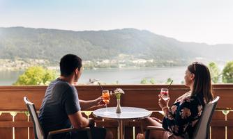 A couple relaxes on a terrace overlooking a lake and the surrounding mountains. They enjoy drinks and the peaceful atmosphere.