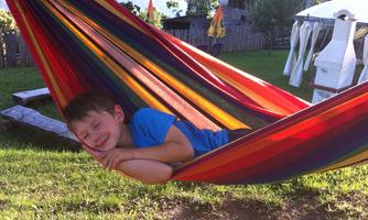 A boy is smiling in a colorful hammock. The background features a green meadow and a few trees.
