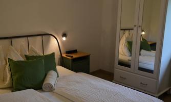 A cozy bedroom with a double bed, decorative pillows, and a mirror. The walls are neutral, and there is a nightstand with a lamp.