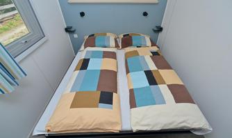 A simple double bed with colorful bed covers in a bright room. The room has a window and simple furnishings.