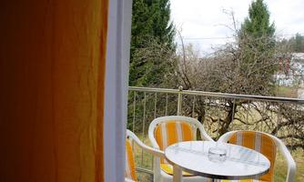 A balcony with two white chairs and a round table. In the background, trees and a cloudy sky can be seen.
