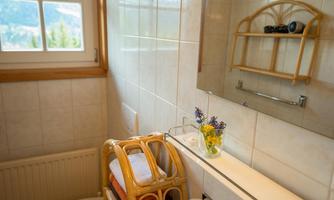 A bright bathroom with a large window and wooden shelves. On the shelf are towels and a small vase with colorful flowers.