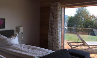 A cozy bedroom with a large window. Outside is a terrace with a lounge chair and a view of the greenery.