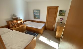 A bright bedroom with two single beds and wooden furniture. Two pictures hang on the wall, and a small wardrobe stands in the corner.