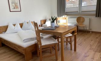 An inviting room with a double bed and a wooden table, surrounded by chairs. Large windows provide plenty of daylight and a view outside.