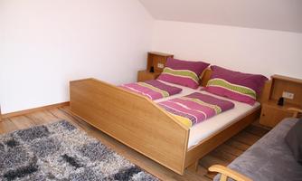 A bright bedroom with a double bed and colorful cushions. The floor is covered with a gray carpet and there is a cozy armchair.