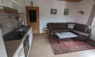 A cozy living room with a dark couch, a table, and a kitchenette. The room features a bright wooden floor and windows with curtains.