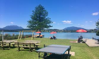 A beautiful lake with a view of the mountains and a green meadow. In the foreground, there is a table tennis table surrounded by loungers and sun umbrellas.