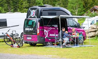 A motorhome with a purple color mark is parked at a campsite. A family member is sitting at the table and enjoying the time outdoors.