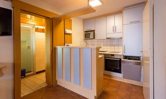A modern kitchen with white cabinets and a cooking area. On the left side of the image, there is a small hallway with access to a bathroom.