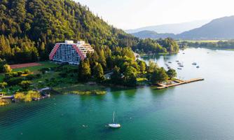 A beautiful lake with green shores and a modern hotel nearby. A few boats can be seen in the water, surrounded by lush nature.