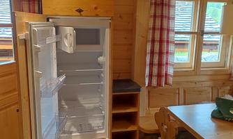 A modern kitchen with an open refrigerator is in the foreground. The room has wooden walls and large windows with checked curtains.
