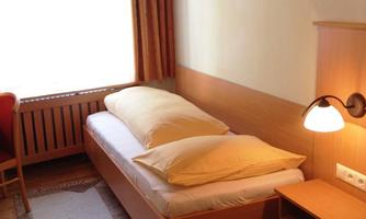 A simple room with a single bed and two pillows. Next to the bed is a nightstand with a lamp.