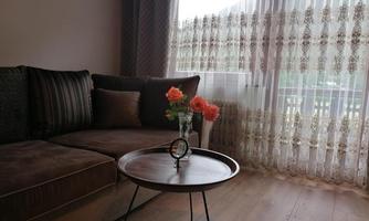 A cozy living room with a brown couch and a round table. On the table sits a vase with pink flowers and large windows with curtains.