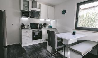 A modern kitchen with white furniture and a cooking island. There are chairs at the table and windows that let in daylight.