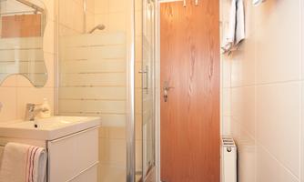 A modern bathroom with a shower and a sink. The walls are tiled in a light color, and the door is made of wood.
