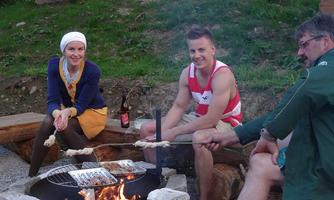 A group of three people is sitting around a campfire and grilling. In the background, green spaces and rocks can be seen.