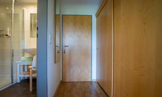 A bright hallway with wood paneling and a closed door. On the left side is a shower and a small table with a chair.