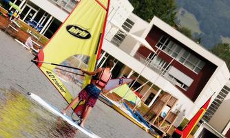 A windsurfer glides over a calm lake, surrounded by modern buildings and mountains. In the background, more windsurfers and colorful sails are visible.