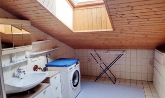 A bright, cozy bathroom with a sloped ceiling and wooden ceiling. It contains a washing machine, a sink, and a clothes drying rack.