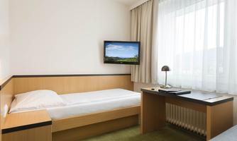 A simple, modern room with a single bed, a desk, and a wall-mounted TV. Large windows let in plenty of daylight.