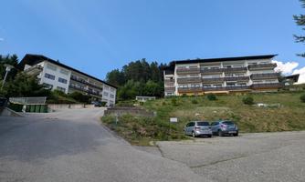 Two residential buildings on a slope, surrounded by trees. The sky is clear and blue.