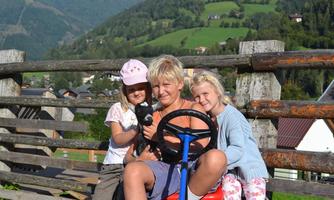 A group of three children is sitting on a toy tractor. In the background, green hills and a rural landscape can be seen.