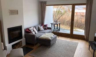 A cozy living room with a gray sofa and a small table. Large windows let in plenty of light and provide a view of the garden.