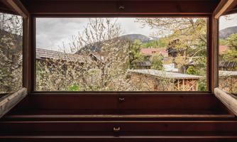 A view through a large window frame that reveals the sight of blooming trees and a green landscape. In the background, rooftops of buildings and gentle hills can be seen.