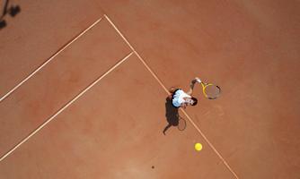 A tennis court viewed from above with a player who is just serving. The court is in a warm terracotta tone, and the player is wearing blue sportswear.