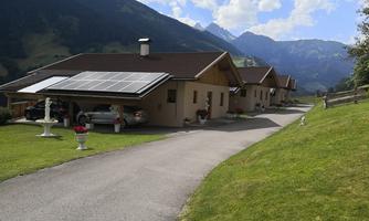 A quiet, rural residential area with modern houses and solar panels on the roof. In the background, green meadows and majestic mountains can be seen.