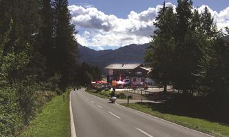 A quiet street, surrounded by trees and mountains. In the background, a building with event tents can be seen.