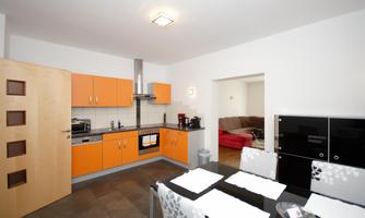 A modern kitchen with orange cabinets and high-quality appliances. In the background, there is a living area with a red sofa.