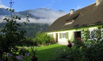 A cozy house with a green garden and colorful flowers. The background features majestic mountains under a clear blue sky.