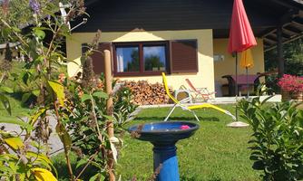 A cozy house with a red sun umbrella and a birdbath in the garden. Some firewood is neatly stacked, surrounded by beautiful plants.