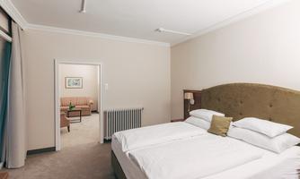 A cozy bedroom with two beds and a large headboard. In the background, a living room with a sofa and a table can be seen.