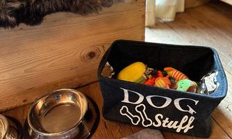 A nice area for a dog with a basket full of toys and two food bowls. The floor is made of wood, and a cozy blanket is available.