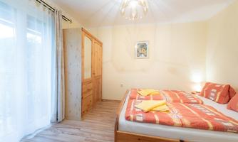 A cozy bedroom with two beds, a wooden wardrobe, and soft colors. The room has large windows that let in plenty of light.