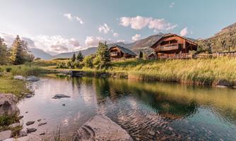 A tranquil landscape with a shimmering lake and green meadows. In the background, cozy wooden houses and mountains stand.