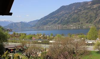 A tranquil landscape with a lake, surrounded by green hills and trees. In the background, there are mountains and a clear blue sky.