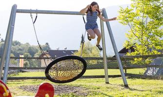 A girl swings joyfully on a large nest swing outdoors. The playground is surrounded by green grass and trees.