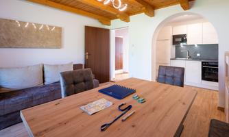 A cozy living space with a wooden table and a modern kitchen in the background. The walls are bright, and there is a relaxed atmosphere.