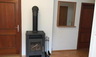 A modern wooden stove stands in a bright room with wooden flooring. In the background, there are two doors and a mirror visible.