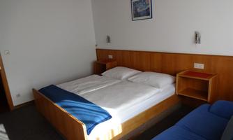 A simple bedroom with a double bed and a blue sofa. The walls are light, and there is a small painting on the wall.