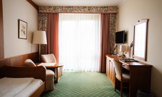 A simple hotel room with a single bed, a table, and two chairs. Large windows let in plenty of daylight.