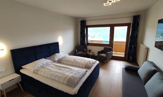 A modern bedroom with a large bed, two chairs, and a table. Large windows offer a view of the outdoor terrace.