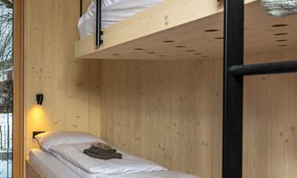 A cozy room with two wooden bunk beds. The walls are made of light wood, and a window lets daylight in.