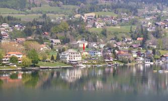 A tranquil landscape with a sparkling lake and beautiful houses along the shore. The hills in the background are green and wooded.
