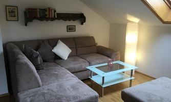 A cozy living room with a gray sofa and a glass table. The light softly filters through a skylight, and a lamp provides additional lighting.