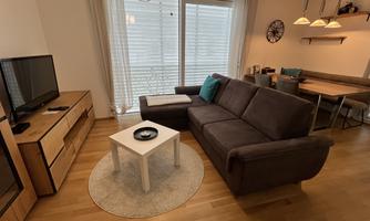 A modern living room with a gray sofa, a small white table, and a TV. Large windows provide plenty of light and create an inviting space.